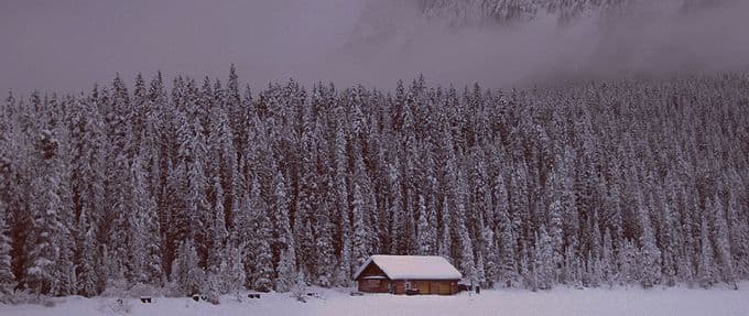 A cabin isolated among trees, covered by feet of snow. Ominous snow clouds hover above the trees
